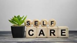 The words SELF and CARE on a table top spelled out with blocks with letters on them. SELF is on top of CARE. Next to the words/blocks there is a small succulent plant. The table is wooden and brown and the wall behind it is grey. 