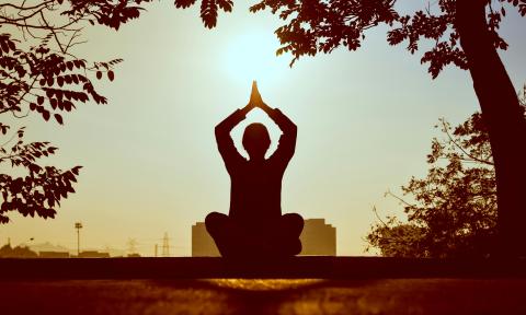 The siloute of a person sitting crosslegged as the sun sets. They have their arms up with their palms touching over their head. 